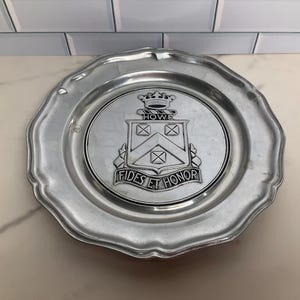 May include: A silver-colored metal plate with a scalloped edge. The center features a crest with the word "HOWE" above a shield design and the words "FIDES ET HONOR" below. The plate is round.