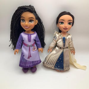 May include: Two dolls standing side by side. The left doll has long black braided hair, a purple dress with a printed belt and tassel, and purple shoes. The right doll has dark brown hair, a cream and blue satin dress with a cape, and green eyes.