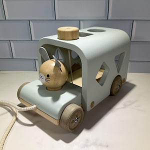 May include: A light blue wooden toy camper with a wooden bunny head. The camper has wooden wheels and a pull string. The bunny head has a painted face and ears. The camper has a wooden top and diamond-shaped cutouts on the sides.