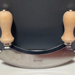 May include: A stainless steel herb chopper with a curved blade and two wooden handles. The handles are light-colored and rounded, attached to the blade with a silver metal collar. The blade has the text "triangle STAINLESS-GERMANY".