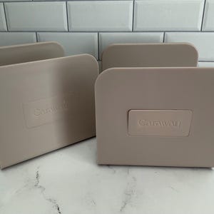 May include: Four beige-colored Caraway brand organizers. Each organizer has a rectangular shape with a curved top and a rectangular label with the brand name. The organizers are made of a solid material and are placed on a white surface.