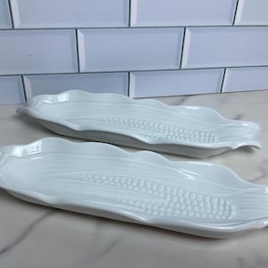May include: Two white ceramic corn-shaped serving dishes. The dishes have a wavy edge and detailed embossed design mimicking corn kernels. They are placed on a white surface, ideal for serving food.