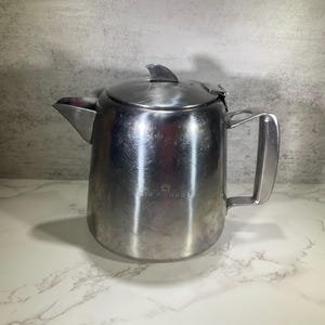 May include: A silver stainless steel teapot with a hinged lid and a handle. The teapot has a logo that says "Aer Products" on the side.