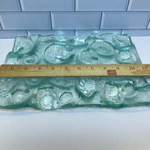 May include: A rectangular, seafoam green glass dish with a textured surface featuring a pattern of swirling shapes and shell-like designs. A wooden ruler is placed across the dish, measuring approximately 12 inches in length.