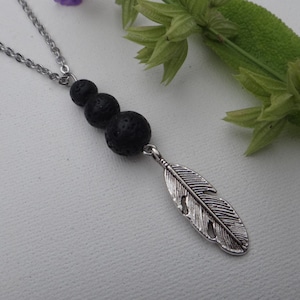 Op de afbeelding: Een zilverkleurige ketting met drie zwarte lavabeads en een gedetailleerde veervormige hanger. De ketting wordt getoond met een takje groene en paarse bloemen, wat een natuurlijke en elegante uitstraling creëert. De ketting is delicaat en de hanger is ingewikkeld ontworpen.