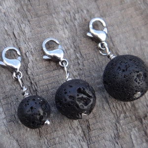 May include: Three black lava rock charms with silver lobster clasps. The spherical lava rock beads have a porous texture. The clasps are attached with silver wire. The charms are displayed on a wooden surface.