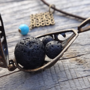 Puede incluir: Un collar con un colgante en forma de jaula de bronce, con una abertura con bisagra, que contiene cuatro cuentas de roca de lava negra. La jaula tiene un diseño delicado y adornado.