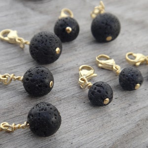 May include: A collection of black lava rock charms with gold-coloured clasps and accents. The charms are spherical and textured, each featuring a small gold bead. The clasps are open, ready to be attached to a bracelet or necklace.