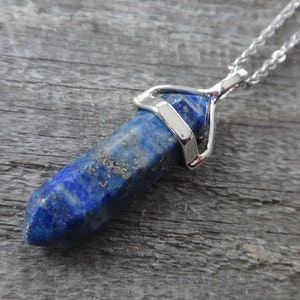 Puede incluir: Un collar de cadena de plata con un colgante de cristal de lapislázuli azul. El cristal está facetado y tiene un engaste de plata.