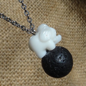 Puede incluir: Un collar de cadena plateada con un dije de elefante de cerámica blanca posado sobre una cuenta de roca de lava negra.