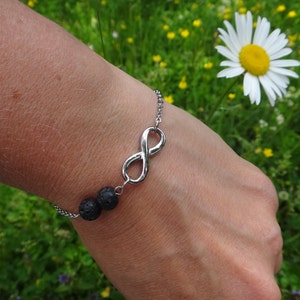 Puede incluir: Un brazalete de plata con un dije de infinito y dos cuentas de lava negra. El brazalete se lleva en la muñeca de una persona sobre un fondo de hierba verde y una margarita blanca.
