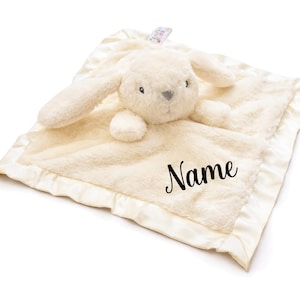 May include: A cream-colored baby security blanket with a plush bunny head and satin trim. The blanket has the word "Name" printed on it in black script. The bunny has long ears and a gray nose.
