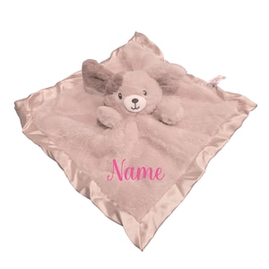 May include: A plush, light gray security blanket with a satin border and a stuffed animal dog head in the center. The dog has gray ears and a light gray body. The word "Name" is embroidered in pink script on the blanket.