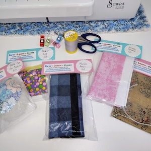 May include: Five different sewing kits with fabric and instructions. The kits include a baby cap, a pocket hug, a mobile mask, a scrunchie, and a sweater cleaner. Each kit has a label with the text "Sew Learn Create" and the name of the kit.