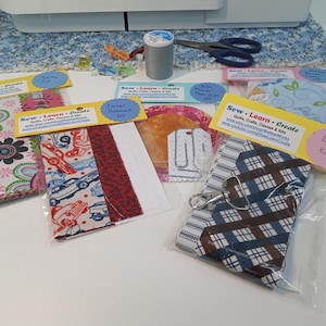 May include: A variety of fabric craft kits, including a car themed kit, a floral kit, a red and white kit, a tag kit, and a lanyard kit. The kits are packaged in clear plastic bags with yellow and white labels that say "Sew Learn Create".