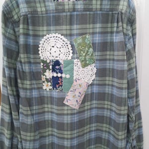 May include: A blue and green plaid flannel shirt with various fabric patches sewn on the back. The patches include floral prints, crocheted doilies, and a green fabric with a leaf pattern.