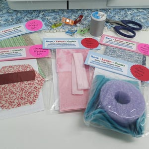 May include: Five different craft kits with pink, blue, and green fabric, and a purple foam ring. The kits are labeled "Sew Learn Create" and include titles like "Lid Gripped Kit", "Hexagon Keychain Kit", "Paw Noodle Cover Kit", and "Bag Handle Cover Kit".