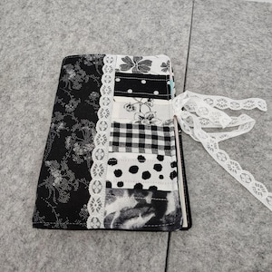 May include: A handmade journal with a black floral patterned cover and a patchwork spine. The spine features various black and white patterns, including polka dots and gingham. A white lace ribbon is tied to the side. The journal is approximately 6 inches tall.