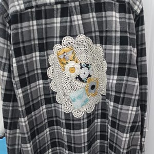 May include: A black and gray plaid flannel shirt with a white crocheted doily and fabric scraps sewn on the back.