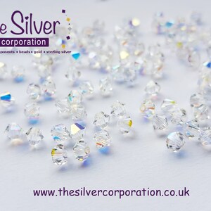 May include: A close-up of a group of clear glass beads with a rainbow iridescent finish. The beads are scattered on a white surface. The beads are small and round, with a faceted design. The beads are perfect for jewelry making.