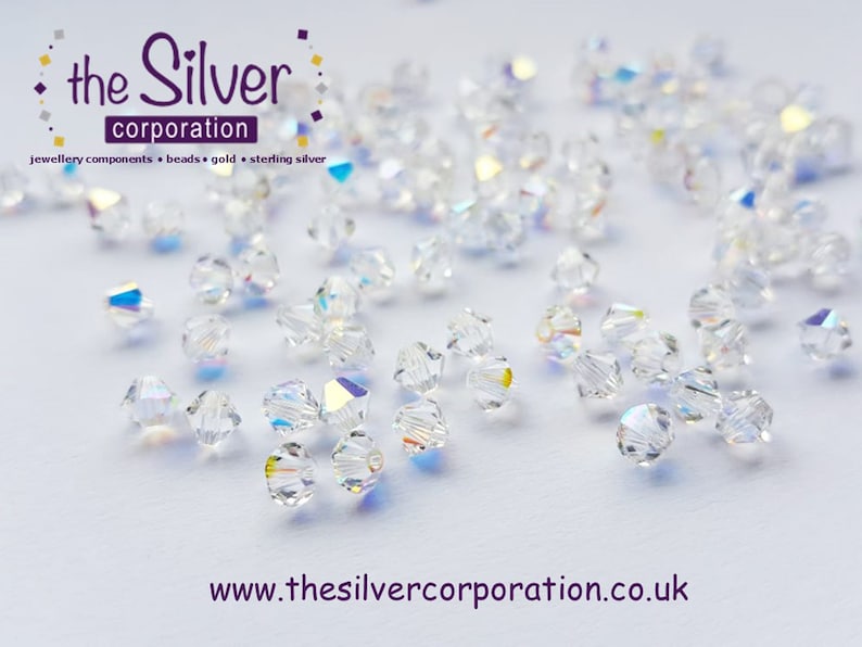 May include: A close-up of a group of clear glass beads with a rainbow iridescent finish. The beads are scattered on a white surface. The beads are small and round, with a faceted design. The beads are perfect for jewelry making.