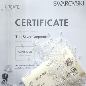 May include: A white certificate with the text "SWAROVSKI CERTIFICATE" and "The Silver Corporation has been certified as a SWAROVSKI AUTHORIZED RETAILER". A bag of clear, iridescent XILION beads is in the lower right corner of the image.