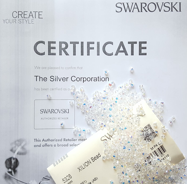 May include: A white certificate with the text "SWAROVSKI CERTIFICATE" and "The Silver Corporation has been certified as a SWAROVSKI AUTHORIZED RETAILER". A bag of clear, iridescent XILION beads is in the lower right corner of the image.