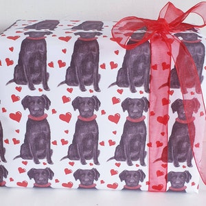 May include: A white gift wrap with a repeating pattern of black Labrador Retrievers wearing red collars and red hearts. The gift is tied with a red sheer ribbon.