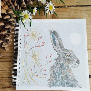 Könnte beinhalten: Ein spiralgebundenes Notizbuch mit einem grauen Einband, der eine handgezeichnete Illustration eines braunen Hasen vor weißem Hintergrund zeigt. Der Hase ist von roten Beeren und gelben Blumen umgeben. Die Illustration erinnert an eine Winterszene.