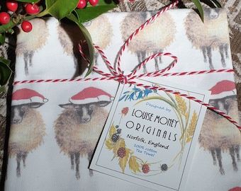 May include: A white tea towel with a pattern of sheep wearing Santa hats. The towel is tied with red and white twine and has a tag that reads "Designed by Louise Money Originals, Norfolk, England, 100% cotton Tea Towel".