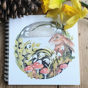 May include: A watercolor illustration of a brown rabbit in a circular frame. The rabbit is looking to the left, surrounded by green leaves, white snowdrops, and red mushrooms.