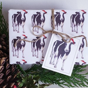 Op de afbeelding: Wit inpakpapier met een herhalend patroon van zwart-witte koeien die kerstmanmutsen dragen. Het papier is opgerold en vastgebonden met een bruin lint. Een cadeaukaart met een zwart-witte koe die een kerstmanmuts draagt, is aan het lint bevestigd.