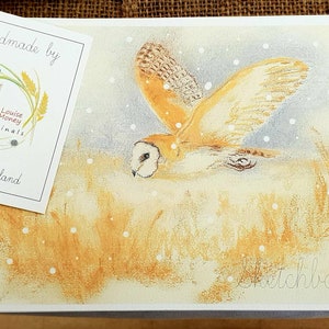 May include: A sketchbook featuring a watercolor painting of a barn owl in flight over a field of tall grass. The owl is depicted with tan and brown feathers, and the scene includes falling snow. The tag reads "Handmade by Louise Money Originals, Norfolk, England."