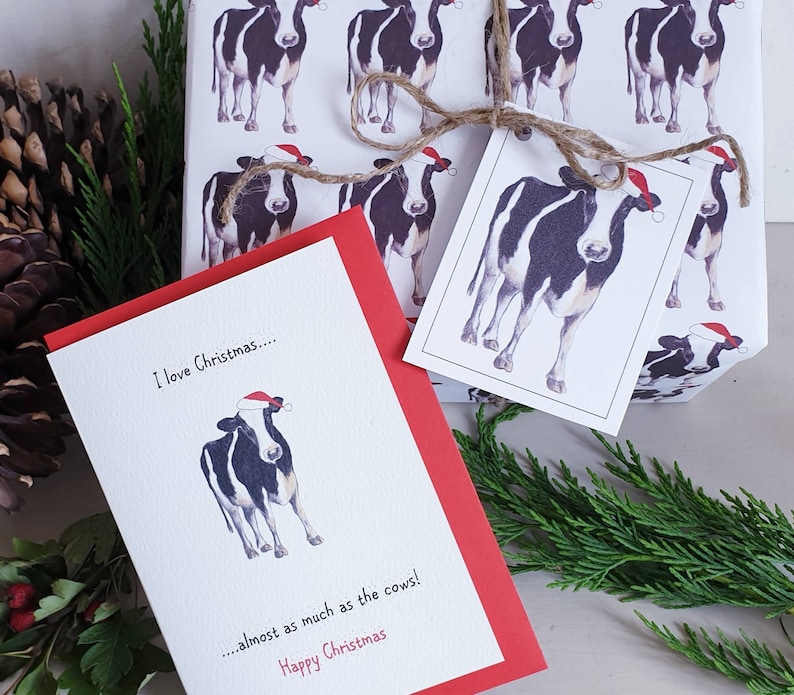 Op de afbeelding: Een rood-witte kerstkaart met een zwart-witte koe die een kerstmanmuts draagt. De kaart staat: "Ik hou van Kerstmis... bijna net zo veel als van de koeien! Fijne Kerst".