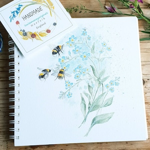 Könnte beinhalten: Ein Aquarellbild eines Straußes Vergissmeinnicht mit drei Hummeln, die um sie herumfliegen. Das Bild ist auf weißem Hintergrund mit einem handgeschriebenen Etikett, auf dem "HANDMADE louisemoneyoriginals NORFOLK England" steht.