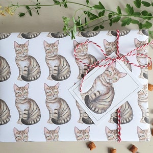 May include: White wrapping paper with a repeating pattern of brown tabby cats. The cats are illustrated in a watercolor style and are facing forward. The paper is decorated with a red and white ribbon.