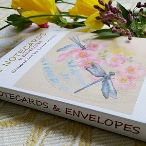 May include: A white box with a light pink and blue watercolour illustration of two dragonflies and flowers. The box contains 8 notecards and envelopes. The text on the box reads "8 Notecards & Envelopes Illustrations by Lovive Money 8 Notecards & Envelopes Printed in Norfolk, England Clovisemoneyoriginals 2016."