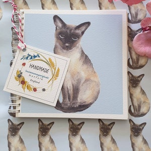 May include: A handmade notebook with a blue cover featuring a watercolour illustration of a Siamese cat. The notebook has a spiral binding and a tag that reads "HANDMADE louisemoneyoriginals Norfolk England".