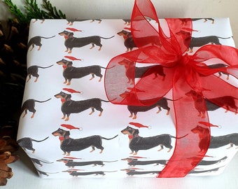 Puede incluir: Papel de regalo blanco con un patrón repetido de dachshunds negros que llevan gorros rojos de Papá Noel. Una cinta roja transparente está atada en un lazo en la parte superior del papel de regalo.