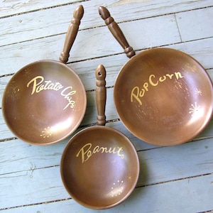 May include: Three brown serving dishes with wooden handles, each inscribed with gold lettering. The dishes are labeled "Potato Chips", "Popcorn", and "Peanut", with starburst designs. The dishes are arranged on a light blue wooden surface.