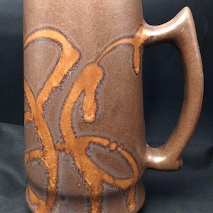 May include: A large, brown ceramic mug with a distinctive handle. The mug features a textured surface and is adorned with abstract, orange-brown decorative patterns. The mug's design suggests it is suitable for beverages.