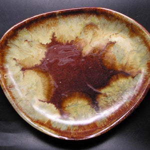 Puede incluir: Un plato de cerámica para servir con una forma orgánica e irregular. El plato presenta una rica paleta de colores terrosos con un centro marrón rojizo intenso, que transiciona a tonos beige cremoso y verde oliva hacia los bordes. El borde es de un tono marrón más oscuro.