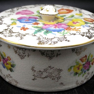 May include: A round, white ceramic trinket box with a lid featuring a cross-stitched floral design in pink, blue, yellow, and green. The box has a gold rim and a gold floral pattern around the sides. A small, gold knob adorns the lid.