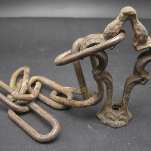 Puede incluir: Una cadena de bronce antigua y un elemento decorativo. La cadena está compuesta por eslabones ovalados, y la pieza decorativa tiene un diseño ornamentado y estilizado. El metal tiene un aspecto desgastado y envejecido, lo que sugiere que es un artículo antiguo.