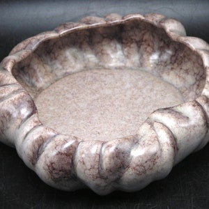 May include: A decorative, round bowl with a textured, speckled interior and a scalloped, rope-like edge. The bowl is primarily beige with brown marbling throughout. The rim has a glossy finish, and the bowl sits on a dark surface.