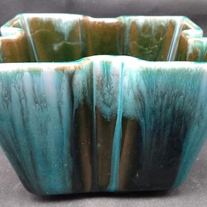May include: A square ceramic planter with a distinctive, wavy rim. The planter has a blend of teal, brown, and white glaze, creating a marbled effect. The interior is a darker shade of green. Suitable for small plants, this is a decorative piece.