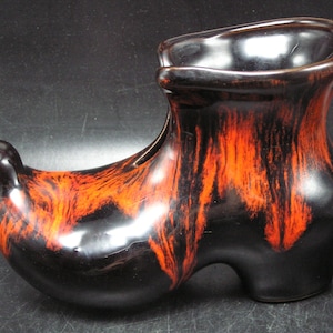 May include: A ceramic shoe-shaped vase with a glossy finish. The vase is mainly black with vibrant orange and red streaks. The shoe has a rounded toe, a curved opening, and a heel. The design is artistic and decorative, suitable for display.