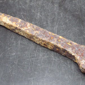 May include: A heavily rusted metal tool, featuring a rectangular shaft and a circular hole at one end. The tool's surface is rough and textured, with a predominantly brown and orange colour. It appears to be made of iron or steel.