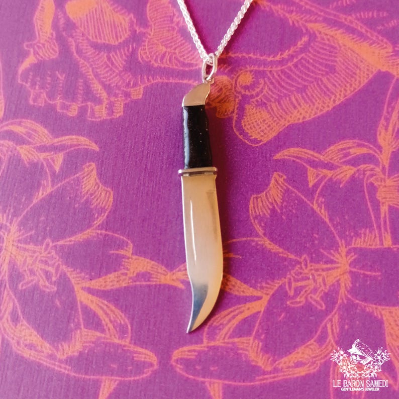 K&ouml;nnte beinhalten: Eine silberne Messer-Anh&auml;nger-Kette mit schwarzem Griff. Der Anh&auml;nger h&auml;ngt an einer silbernen Kette vor einem violetten Hintergrund mit orangefarbenen Blumendesigns.