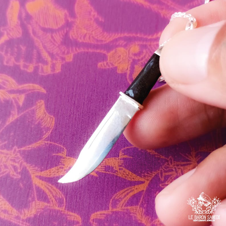 K&ouml;nnte beinhalten: Ein silberner Messeranh&auml;nger mit schwarzem Griff. Das Messer ist etwa 7,5 cm lang und hat eine scharfe Spitze. Der Anh&auml;nger h&auml;ngt an einer silbernen Kette.